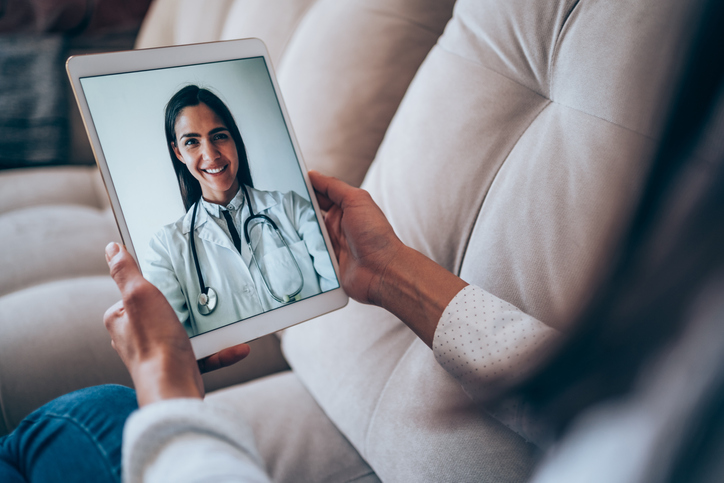 Shot of a patient talking to a doctor using digital tablet. Sick young woman during video call with her doctor. Back view of patient talking during online video conference call with her family doctor while staying at home in quarantine. Young woman in online consultation with general practitioner on digital tablet. Online telemedicine concept.